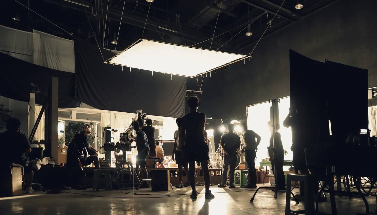 Silhouetted film crew with cameras and lights working on a brightly lit indoor set at night under a large overhead light panel Silhouetted film crew with cameras and lights working on a brightly lit indoor set at night under a large overhead light panel