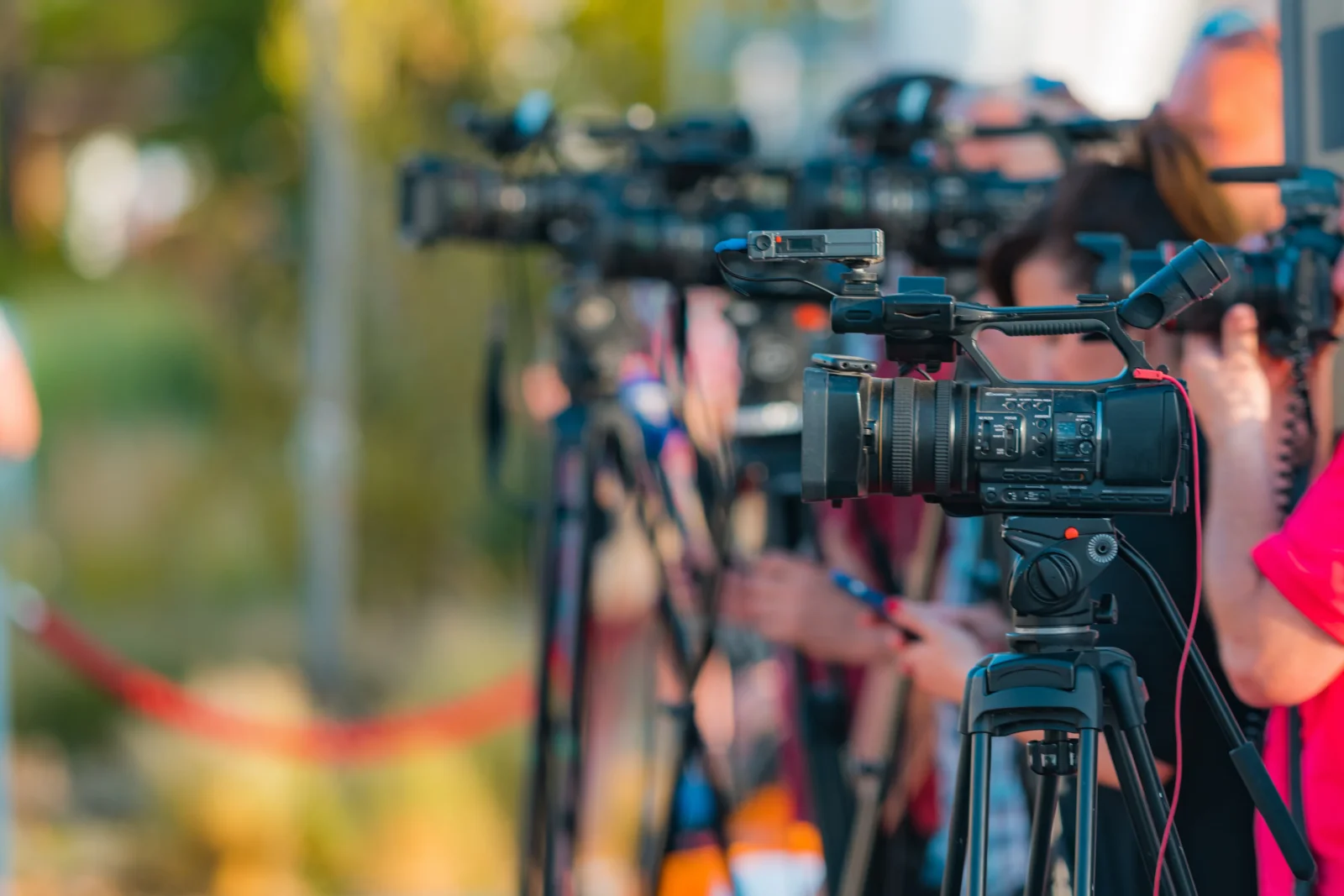 A row of professional news video cameras on tripods aimed at a press conference event outdoors.