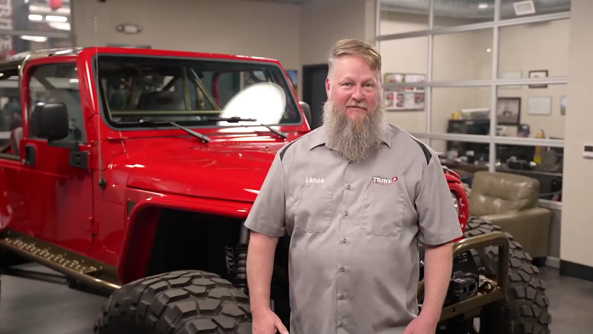 A local business owner on camera A bearded man in a grey work shirt standing in an auto shop in front of a lifted red Jeep, lit for camera during a Sierra shoot.