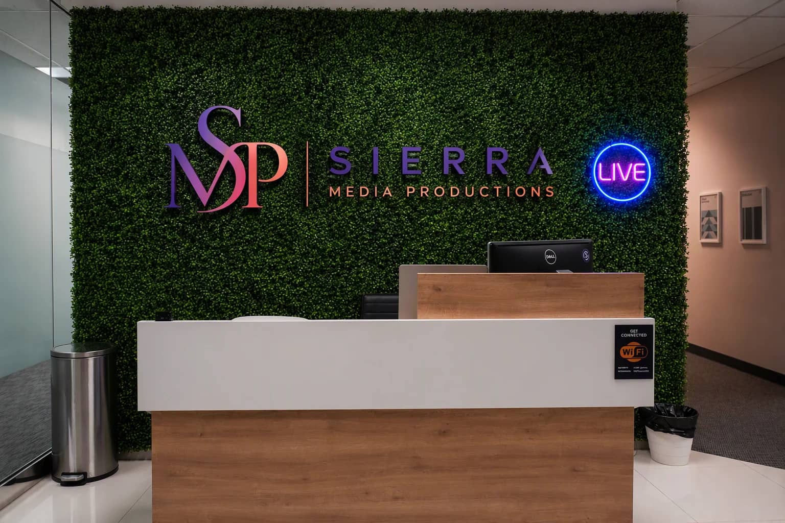 The reception desk and living-greenery wall inside the Dallas podcast studio, with the Sierra Media Productions mark and a LIVE neon sign. The reception desk and living-greenery wall inside the Dallas podcast studio, with the Sierra Media Productions mark and a LIVE neon sign.