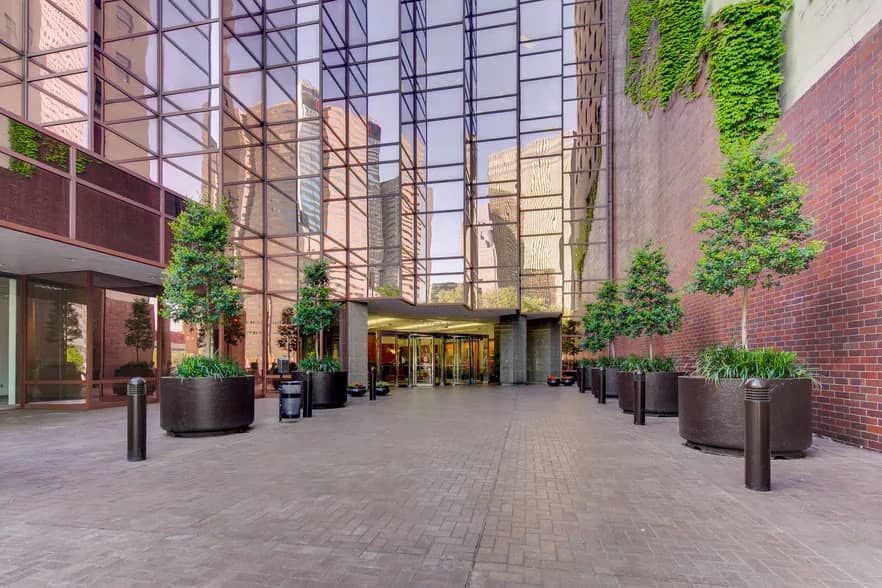 The glass atrium entrance of the downtown Dallas building where Sierra Media Productions records podcasts, with an ivy-covered brick wall and planted trees along the paved plaza. The glass atrium entrance of the downtown Dallas building where Sierra Media Productions records podcasts, with an ivy-covered brick wall and planted trees along the paved plaza.