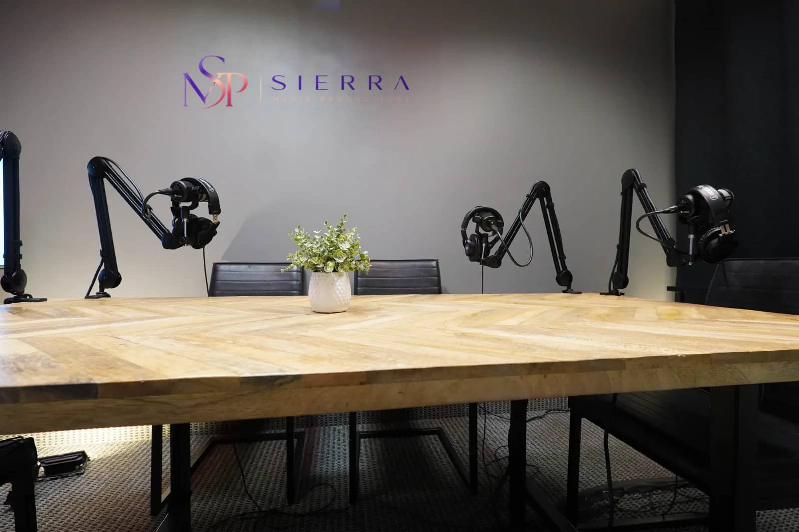 Dallas-Fort Worth podcast recording room A Dallas-Fort Worth podcast recording room: a chevron wood table with four broadcast microphones on boom arms, studio lighting, and the Sierra Media Productions wall mark behind it.