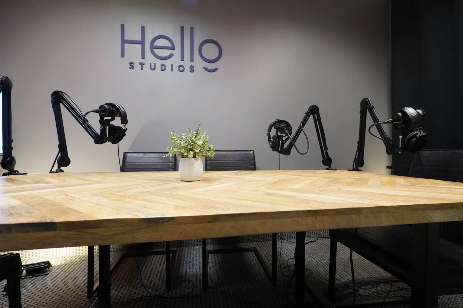 Dallas-Fort Worth podcast recording room A Dallas-Fort Worth podcast recording room: a chevron wood table with four broadcast microphones on boom arms, studio lighting, and the Hello Studios wall mark behind it.