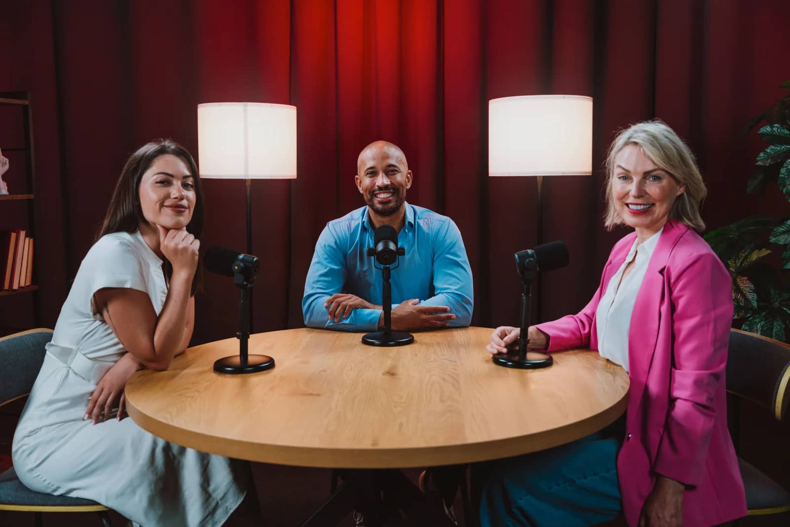 Three smiling people seated at a round wooden table with microphones in a studio with red curtains and floor lamps Three smiling people seated at a round wooden table with microphones in a studio with red curtains and floor lamps