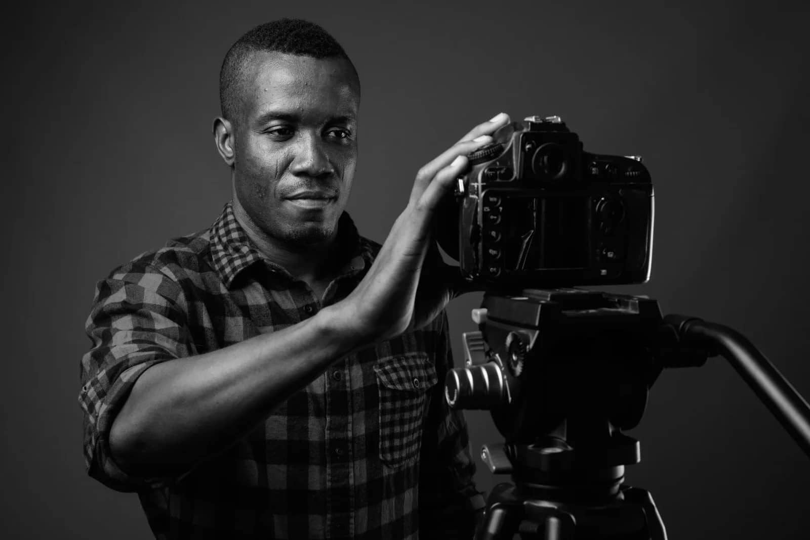 Black-and-white photo of a man in a plaid shirt adjusting a camera mounted on a tripod against a dark backdrop Black-and-white photo of a man in a plaid shirt adjusting a camera mounted on a tripod against a dark backdrop
