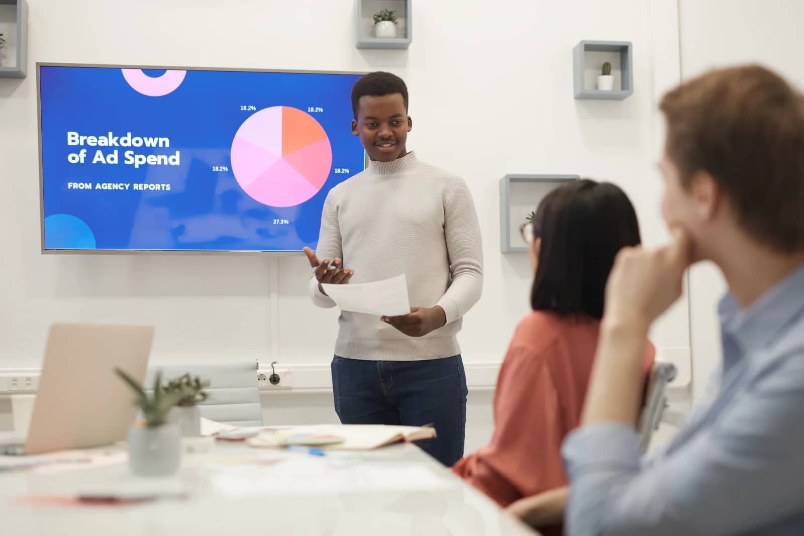 A man presents a screen titled 'Breakdown of Ad Spend' with a pie chart to two seated colleagues in a bright meeting room.