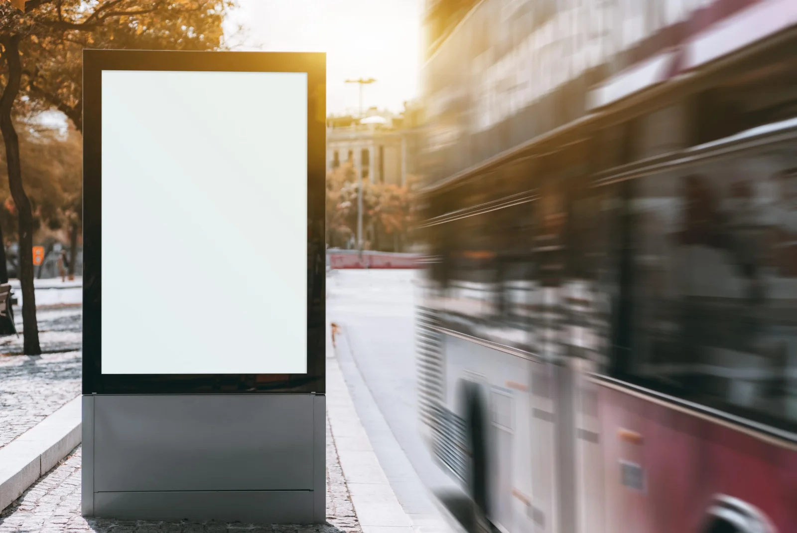 A blank street-side advertising billboard on a city pavement in warm evening light, with a bus rushing past in motion blur