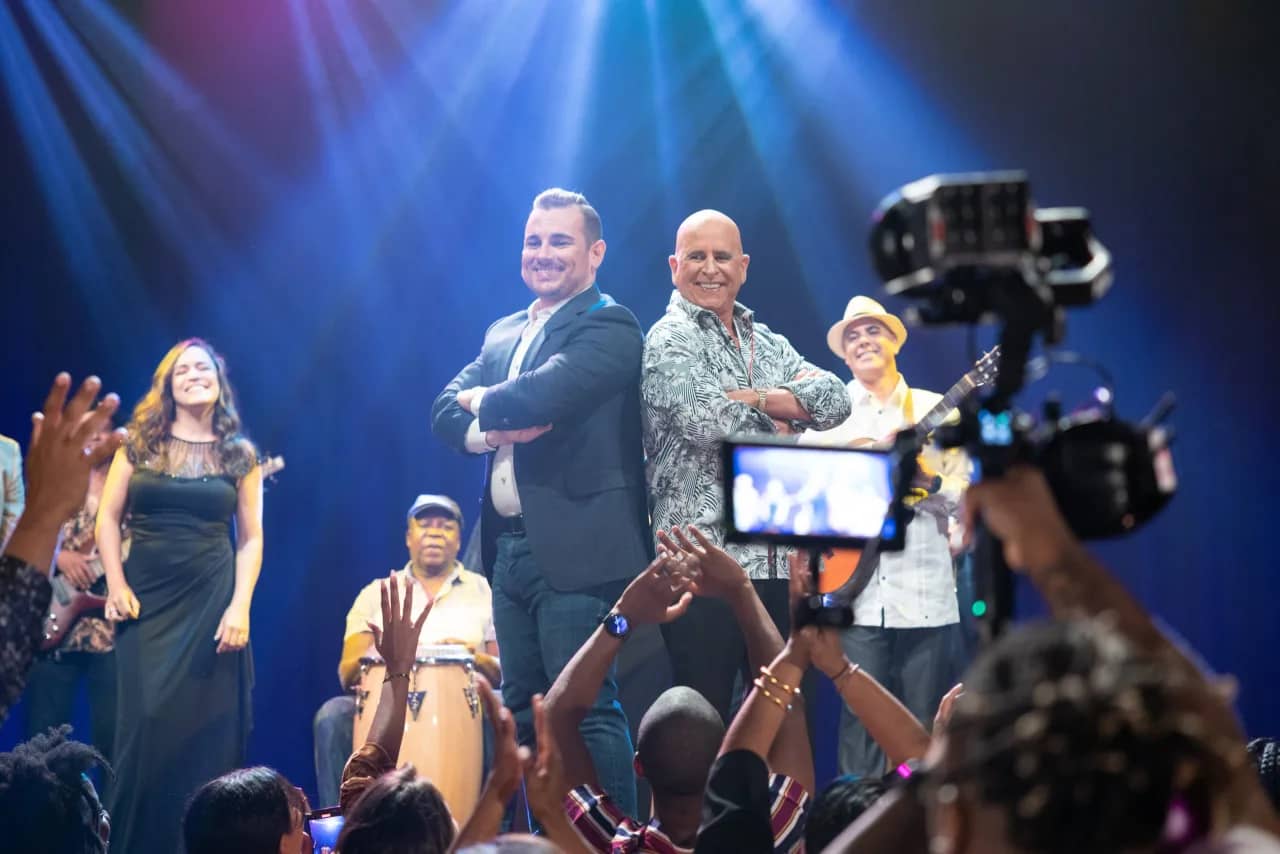 Live event filming A camera operator films two hosts on a blue-lit concert stage while the audience raises their hands at a Sierra Media Productions live event.