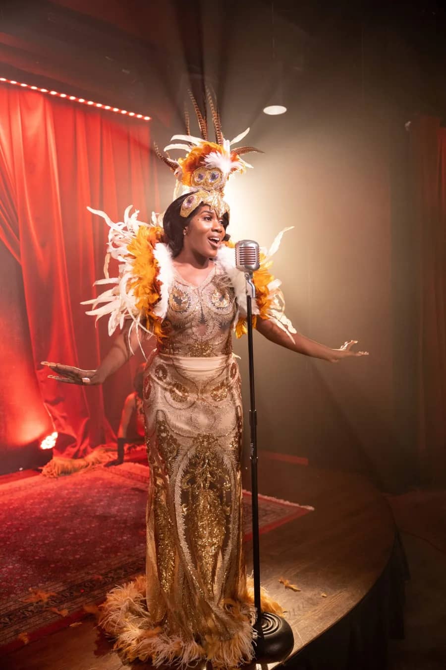 Branded film, stage performance A performer in an elaborate gold feathered costume sings into a vintage microphone on a red-curtained stage in a Sierra Media production.