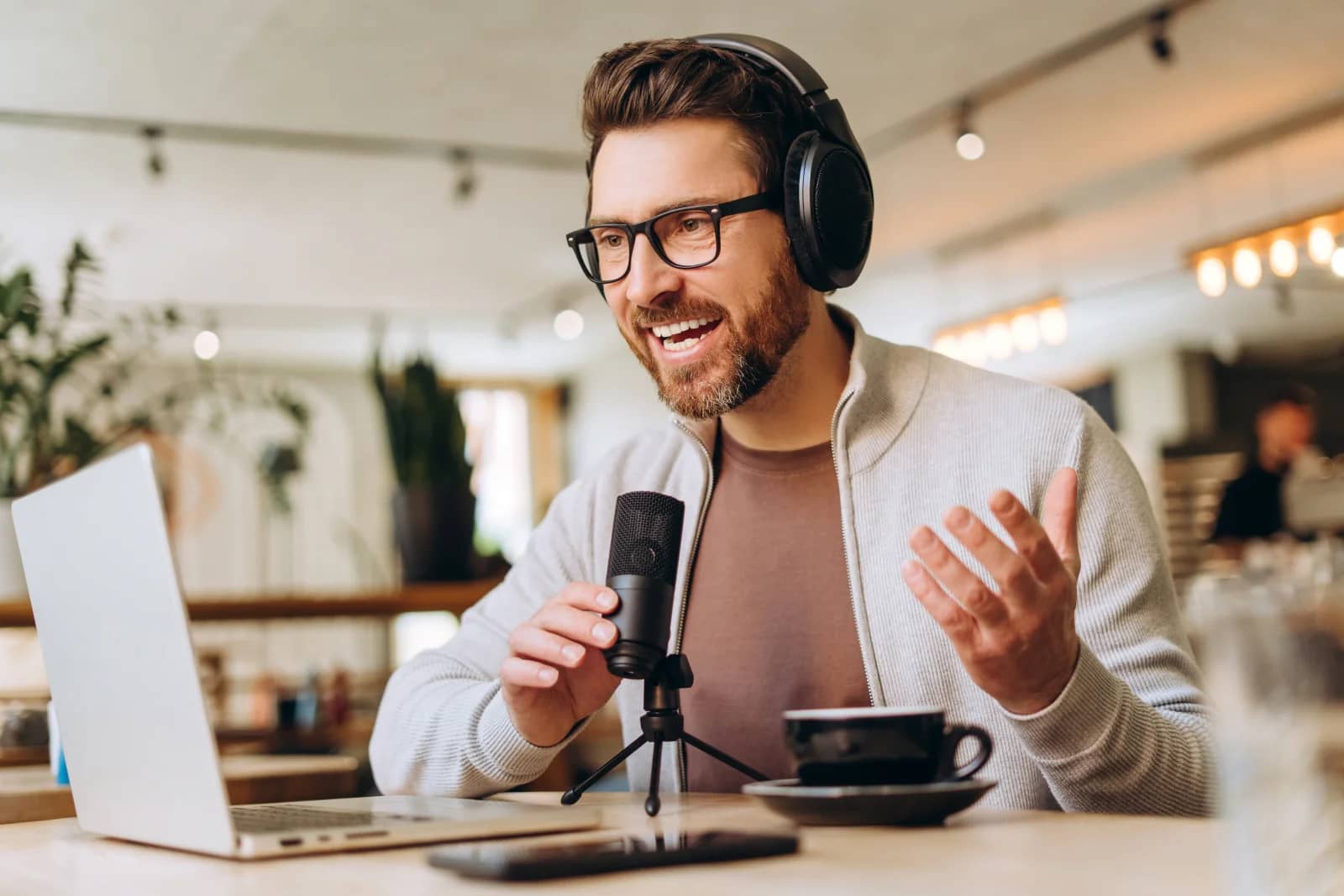 Man in glasses and headphones smiling and speaking into a podcast microphone at a table with a laptop and coffee Man in glasses and headphones smiling and speaking into a podcast microphone at a table with a laptop and coffee