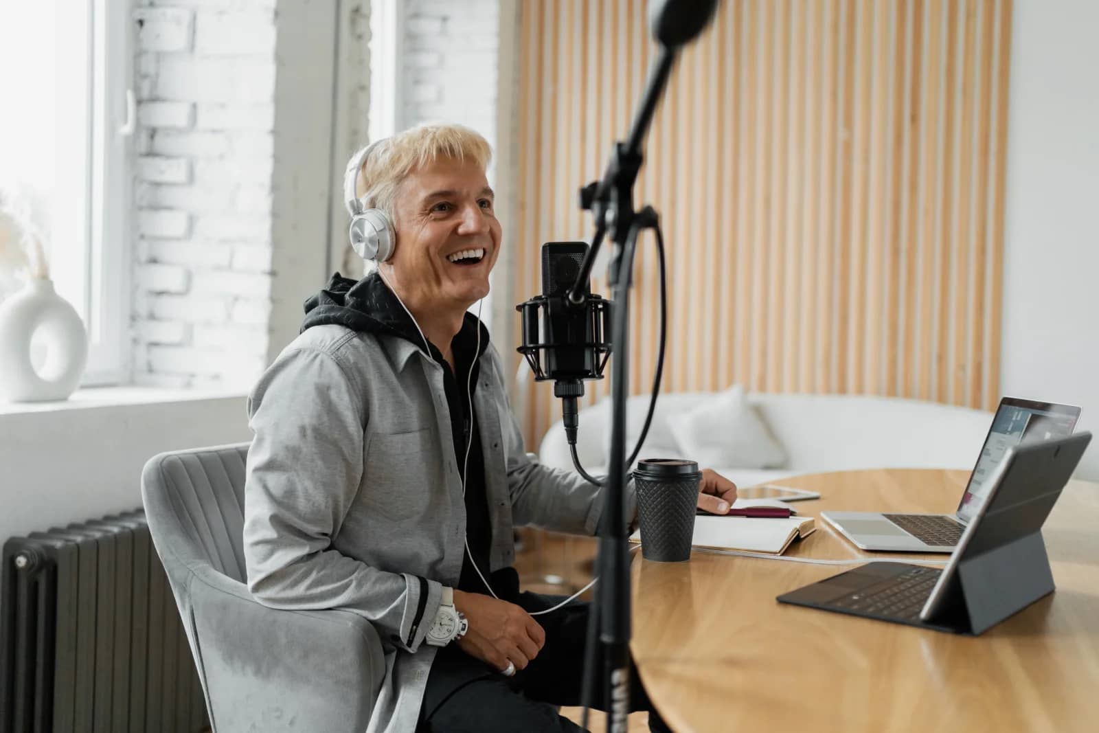 Smiling grey-haired man wearing headphones speaks into a studio microphone at a wooden desk with a laptop and tablet Smiling grey-haired man wearing headphones speaks into a studio microphone at a wooden desk with a laptop and tablet