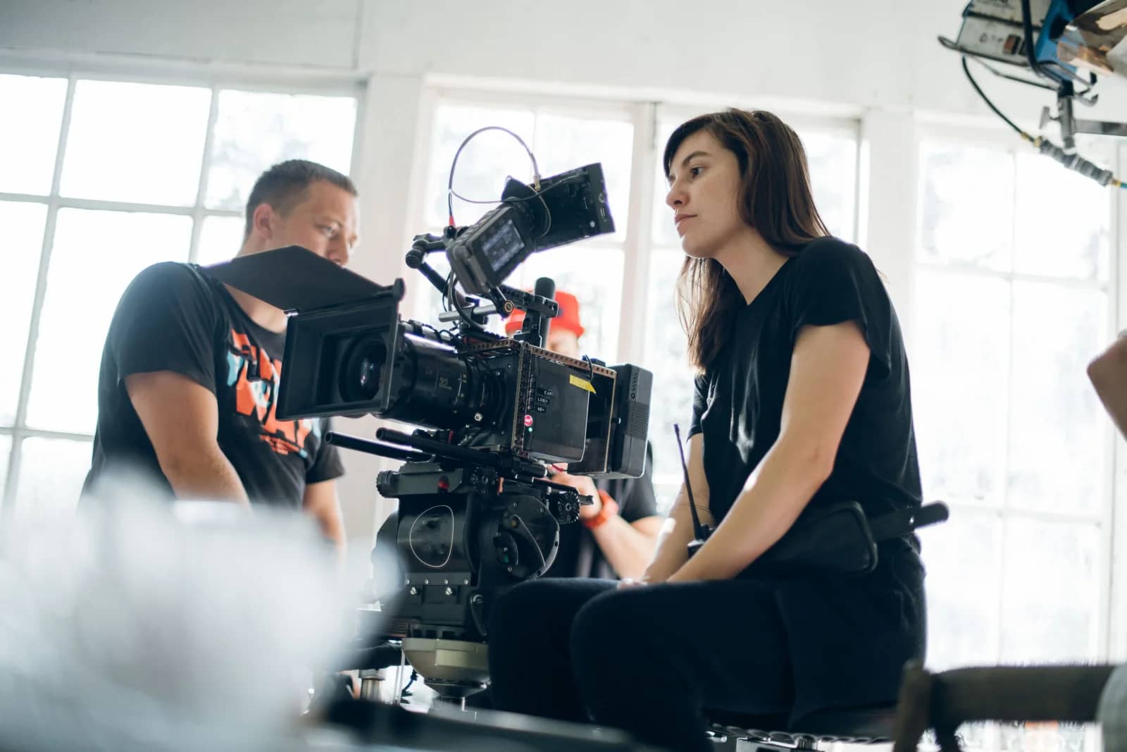 Two film crew members operate a cinema camera in a bright, window-lit studio room. Two film crew members operate a cinema camera in a bright, window-lit studio room.